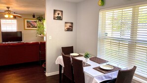 Un coin repas avec une table dressée pour deux personnes, un téléviseur en arrière-plan et une fenêtre avec des stores.