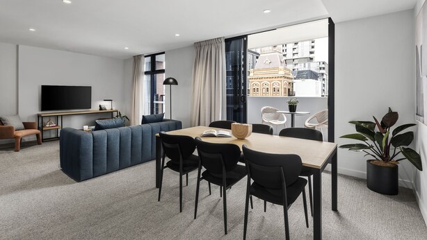 Ein modernes Wohnzimmer mit einer Couch, einem Esstisch und Stühlen. Ein Fernseher, eine Pflanze und ein Blick auf Gebäude durch große Fenster sind zu sehen.