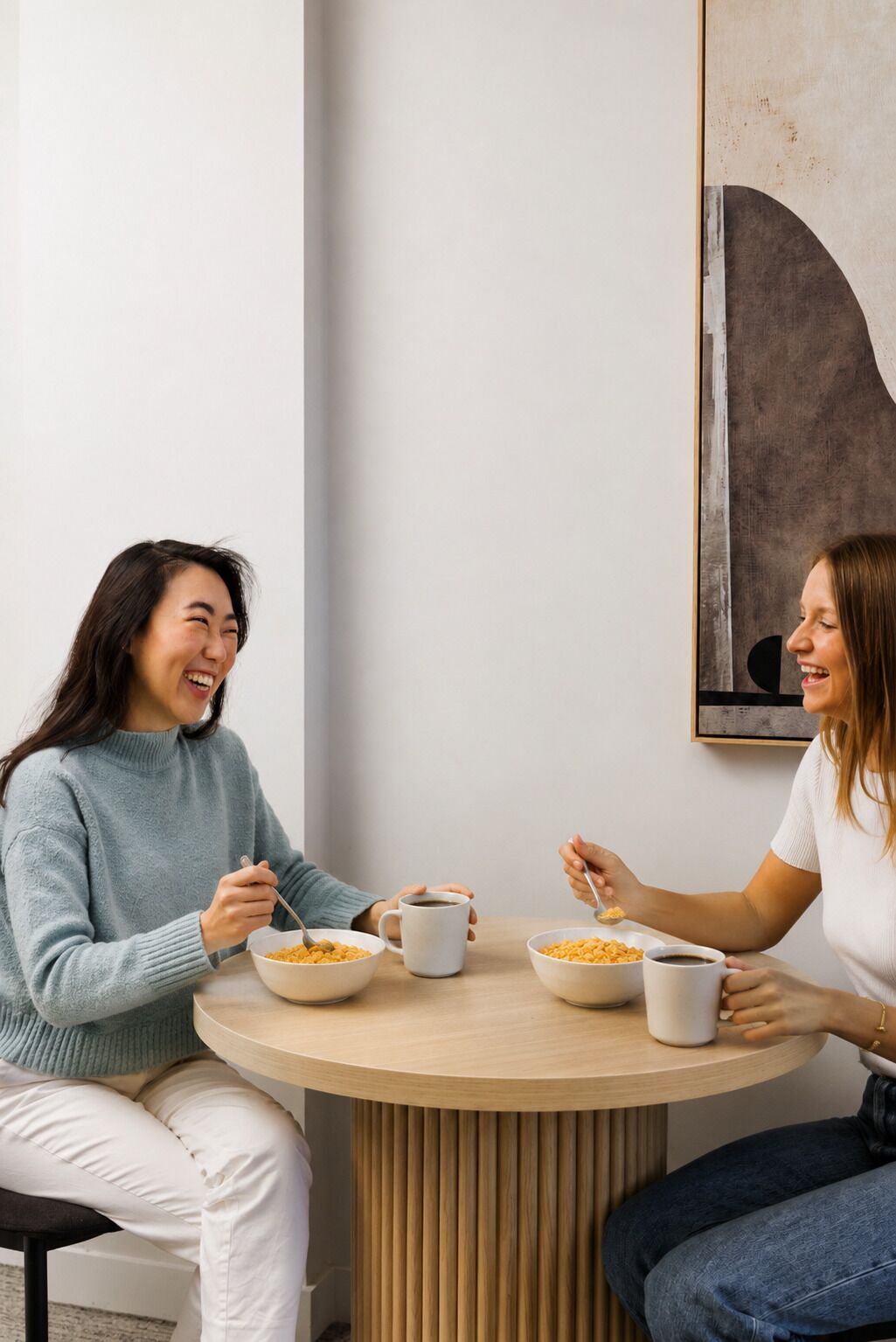 Dos personas sentadas en una mesa redonda de madera, con tazones de cereal y tazas de café.