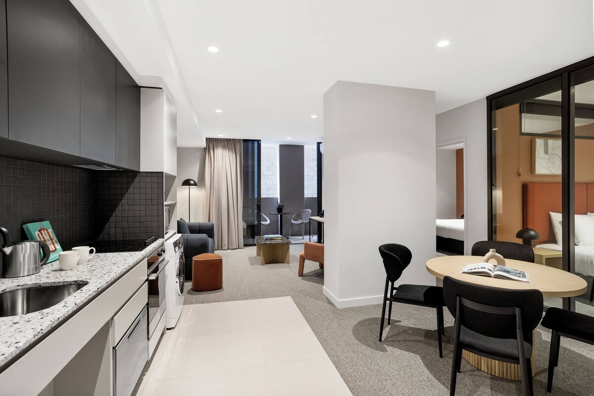 A modern kitchen with a built-in oven, a sink, and a coffee maker. There is a dining area with a round table and chairs.