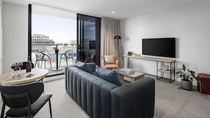 Ein modernes Hotelzimmer mit Sofa, Esstisch, Fernseher und einem Balkon mit Blick auf die Stadt.