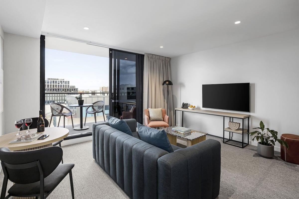 A modern hotel room with a sofa, a dining table, a TV, and a balcony with a city view.