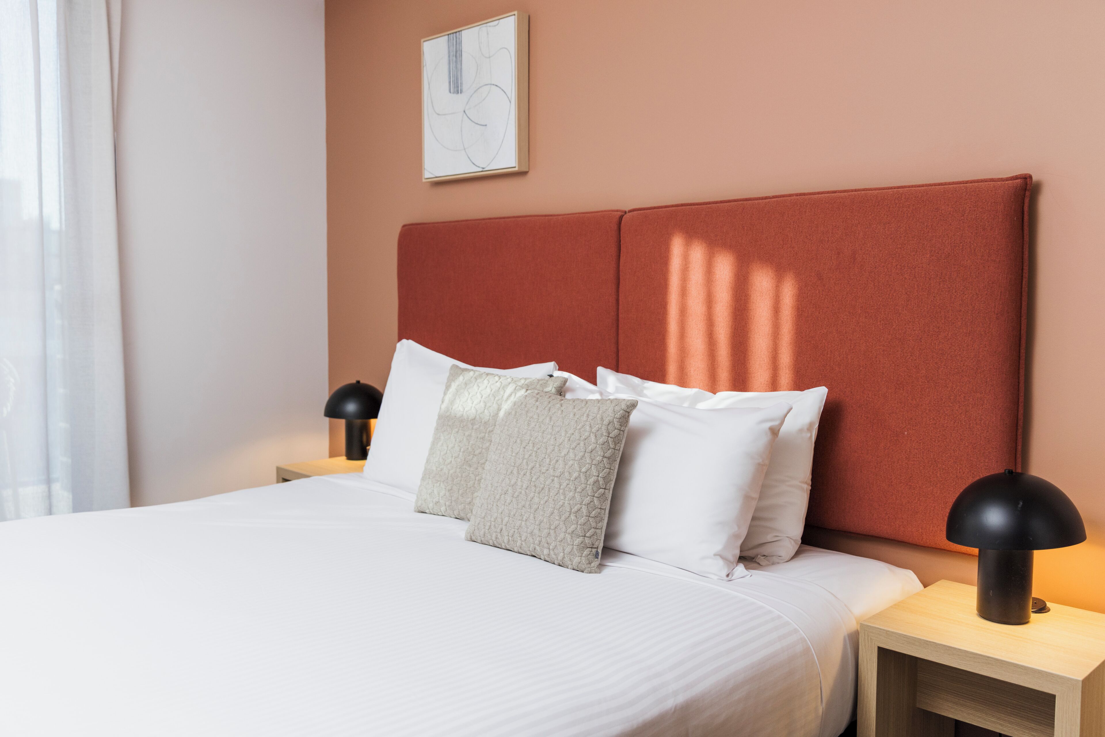 A neatly made bed with white linens and a textured pillow, a bedside lamp, and a framed abstract artwork on the wall.