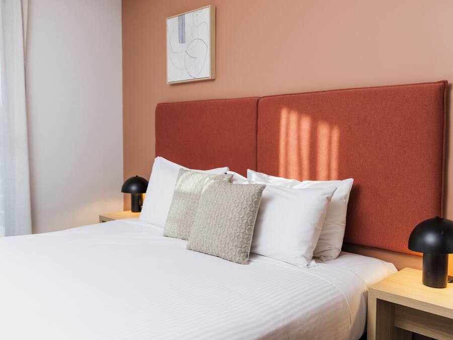 A neatly made bed with white linens and a textured pillow, a bedside lamp, and a framed abstract artwork on the wall.