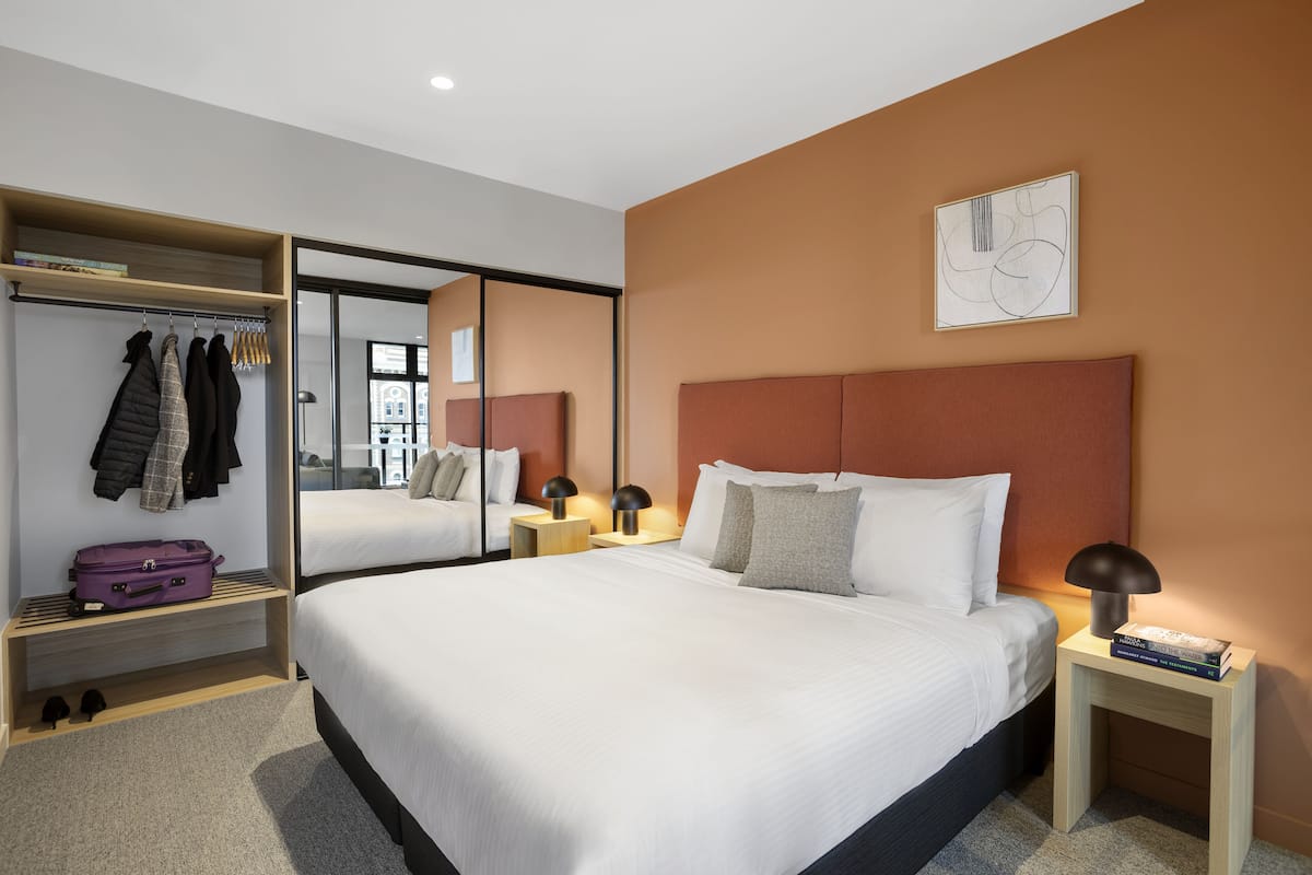 A modern hotel room with a large bed, a wardrobe, a mirror, and a bedside table with a lamp.