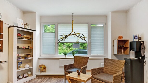 Una habitación con un amplio ventanal, sillas de mimbre, una mesa de madera y una estantería.