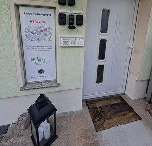 A door with a 'Welcome' mat, a sign with multilingual greetings, and a black lantern.