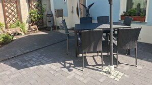 Eine Terrasse im Freien mit Tisch, Stühlen und einem blauen Sonnenschirm, sowie einer Gesichtsstatue.