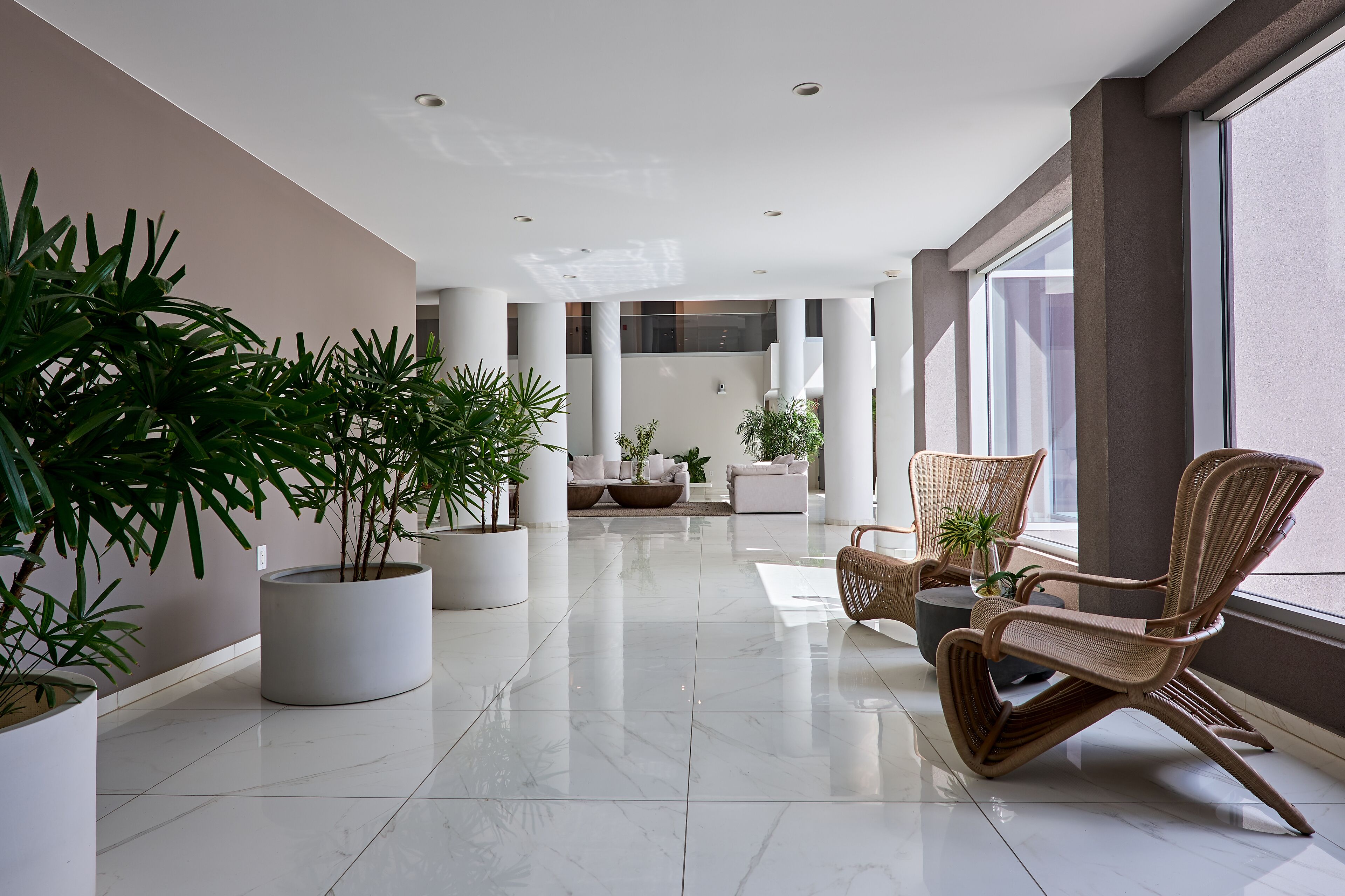 A spacious lobby with a modern design, featuring a seating area with wicker chairs, large potted plants, and a sleek, tiled floor.