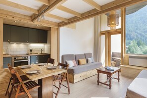 Una cocina moderna con vigas de madera, un área de comedor con mesa y sillas de madera, un sofá y un ventanal con vistas a la montaña.