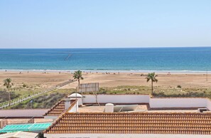 Vista de una playa desde una terraza en la azotea, con techo de tejas, una palmera y una playa de arena al fondo.