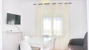 Una habitación moderna con una mesa de vidrio, sillas blancas, un televisor de pantalla plana y un amplio ventanal con cortinas translúcidas.