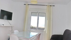 Una habitación con una mesa de vidrio, sillas blancas, un sofá negro, un televisor montado en la pared y una ventana con vista a la playa.