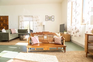 A living room with a wooden sofa, a green couch, and a wooden coffee table. There is a large window with curtains, a floor lamp, and a television on a wooden stand.