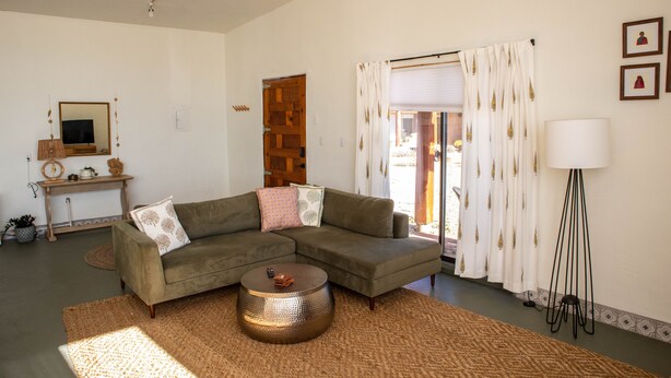 A living room with a sectional sofa, a round coffee table, a lamp, and a view through a window with curtains. - Eclectic, Art-filled Studio with Fantastic Views (El Prado)