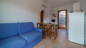 Un espacio habitable pequeño con un sofá azul, una mesa de comedor de madera con sillas, un refrigerador blanco y una pared de piedra visible a través de una puerta.