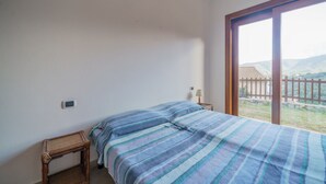 Un dormitorio con cama, mesita de noche, lámpara y un ventanal con vistas a una cerca y montañas.