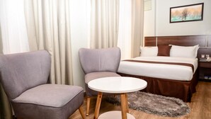 Una habitación de hotel con una cama, dos sillones, una mesita redonda y un cuadro en la pared.