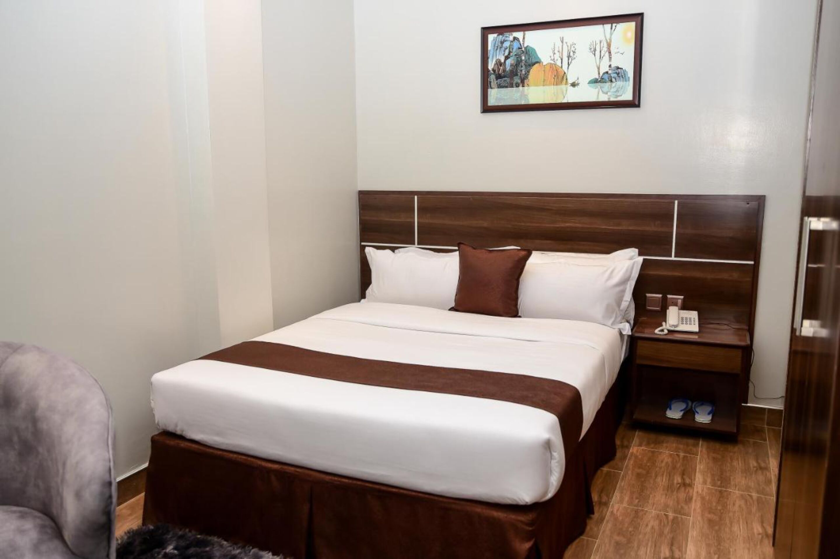 A neatly made bed with a brown headboard, a bedside table with a phone and slippers, and a painting on the wall.