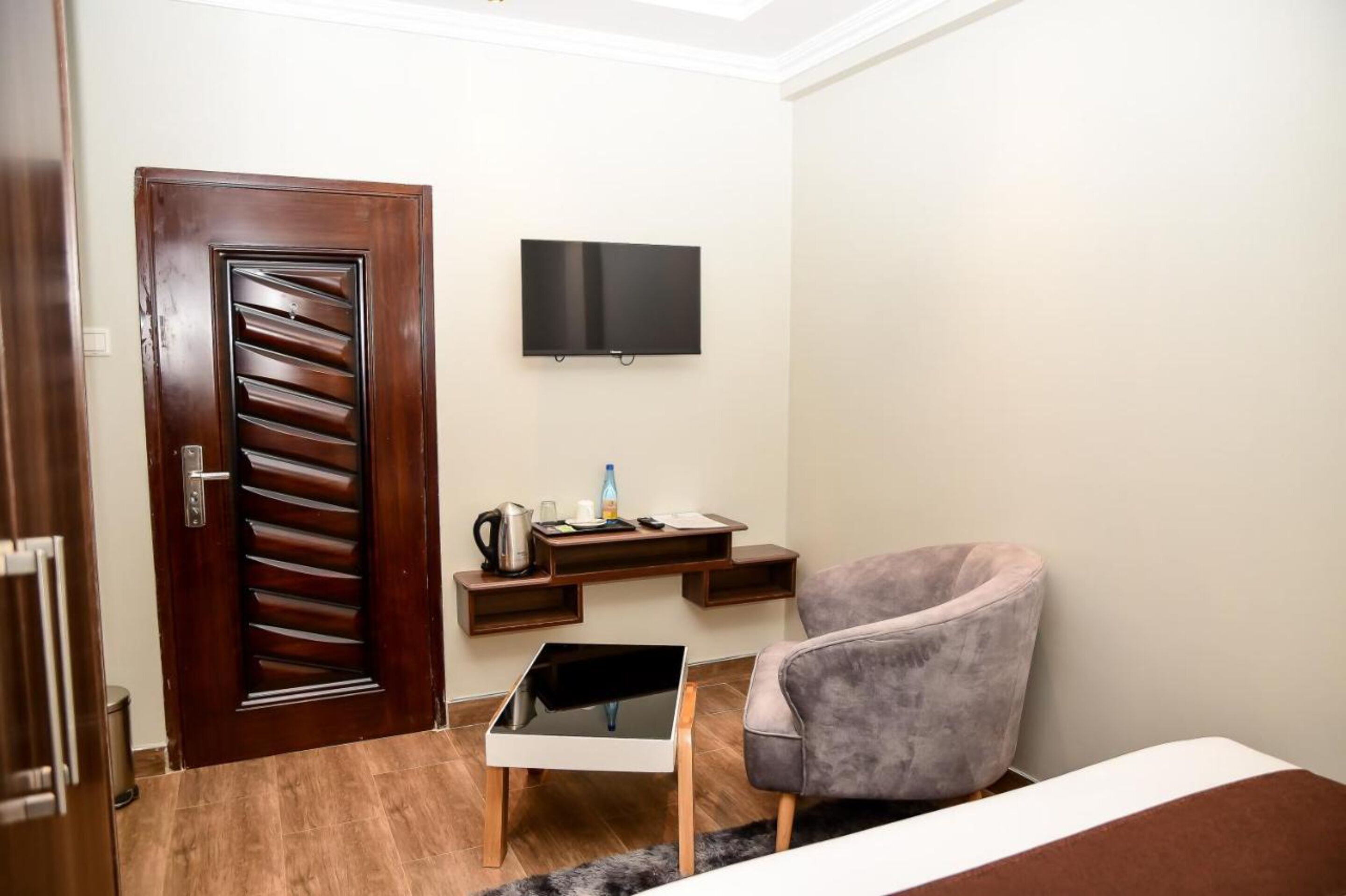 A room with a wooden door, a flat-screen TV mounted on the wall, a small table with a kettle, a chair, and a bed.