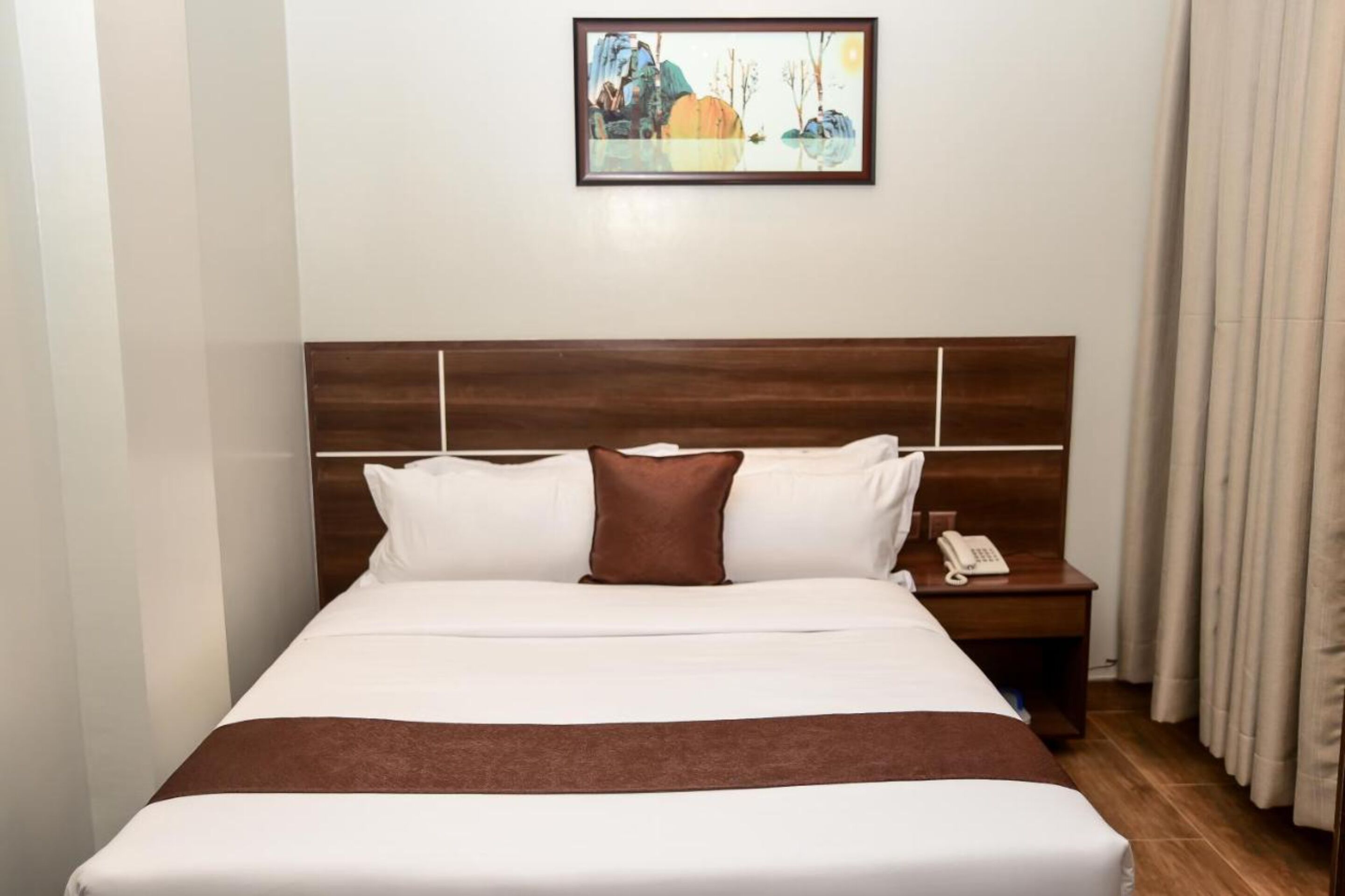 A neatly made bed with white and brown bedding, a wooden headboard, a bedside table with a phone, and a painting on the wall.