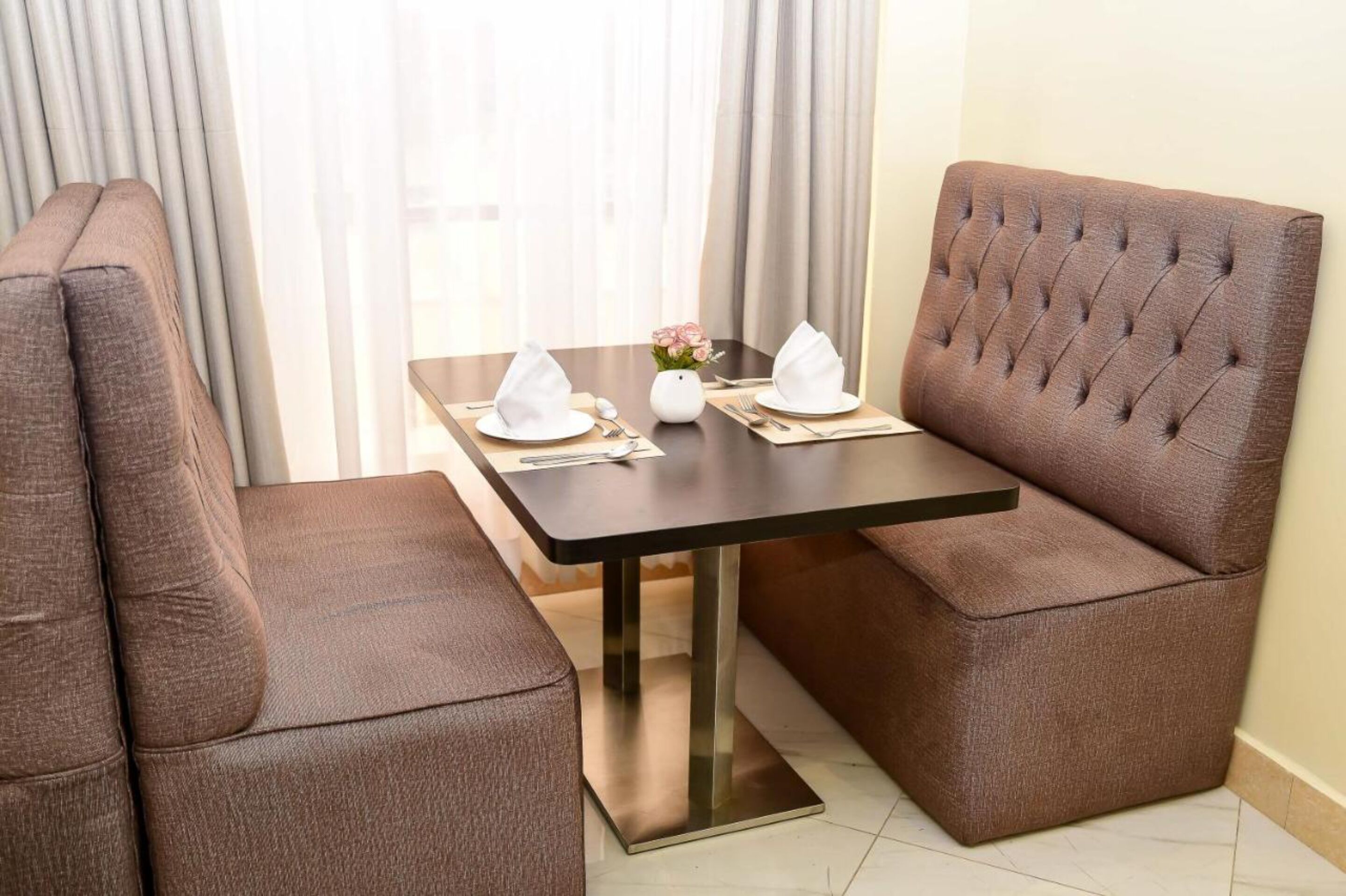 Comedor con una mesa dispuesta para dos personas, con sillas tapizadas en tono marrón y una mesa negra con base metálica.
