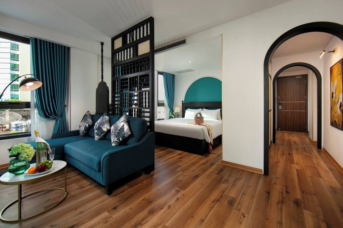 A modern hotel room with a blue sofa, a bed, a bookshelf, and a dining table with fruits and a bottle.