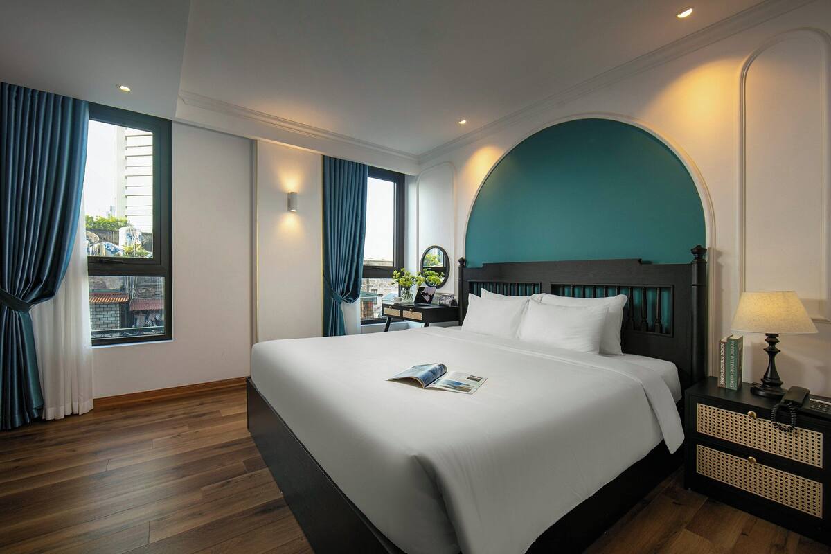 A hotel room with a large bed, a nightstand with a lamp, a window with curtains, and a view of the city.