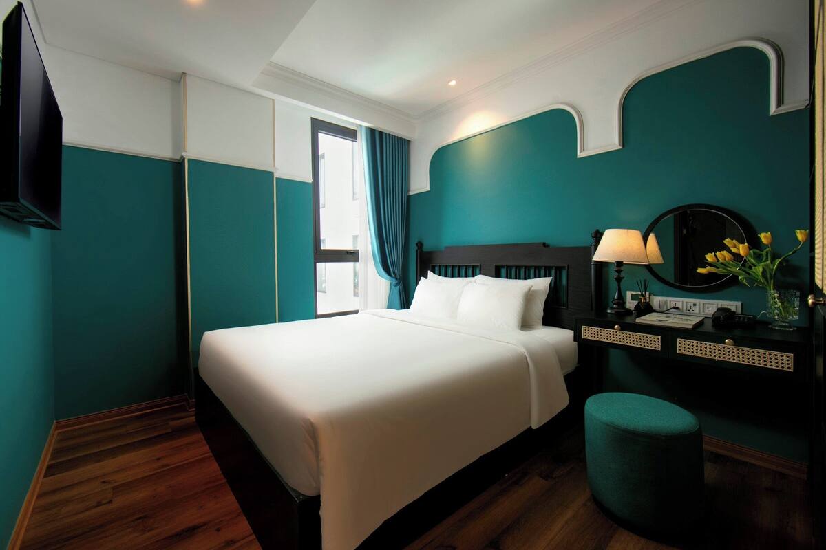 A hotel room with a large bed, a desk with a lamp, a mirror, and a vase with flowers.