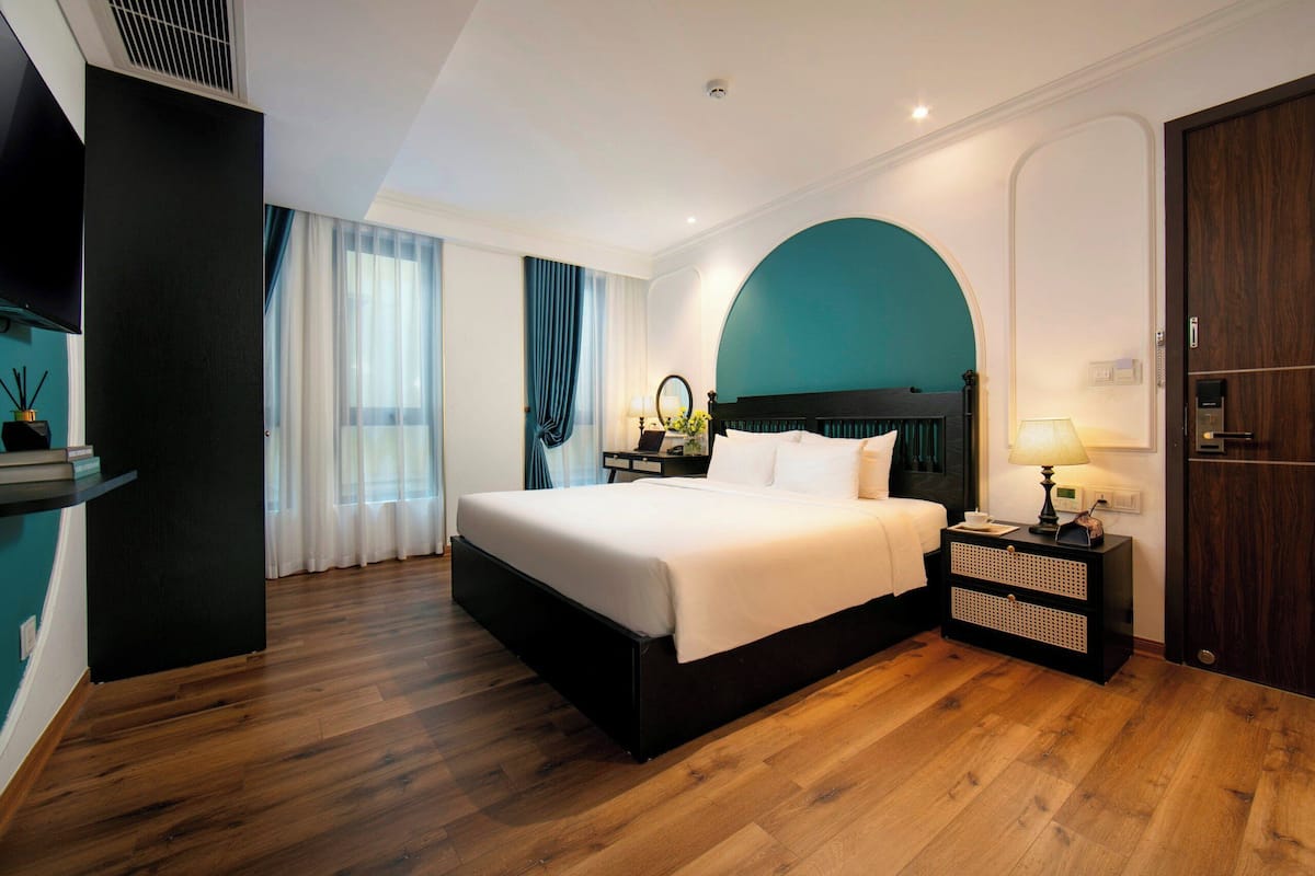 A modern hotel room with a large bed, wooden flooring, a teal accent wall, a nightstand with a lamp, and a TV mounted on the wall.