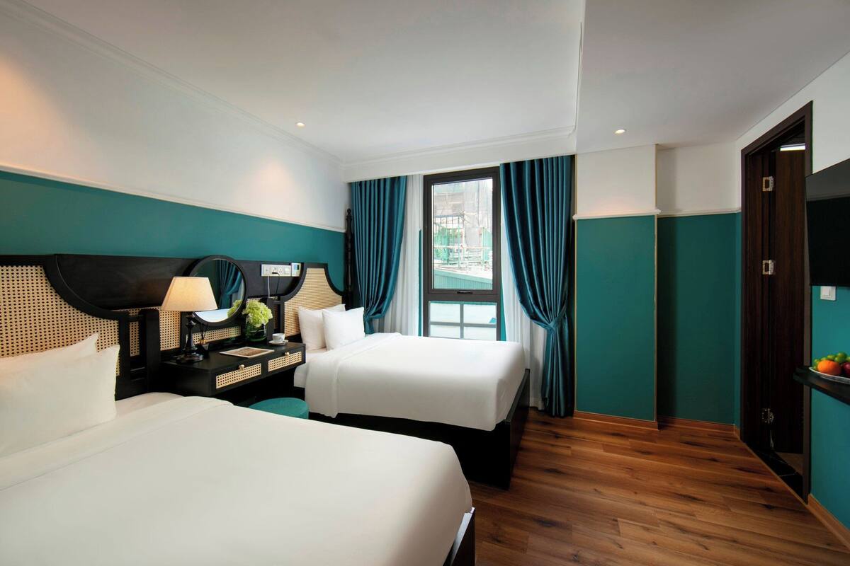 A hotel room with two beds, a desk with a lamp, a window with curtains, and a door.