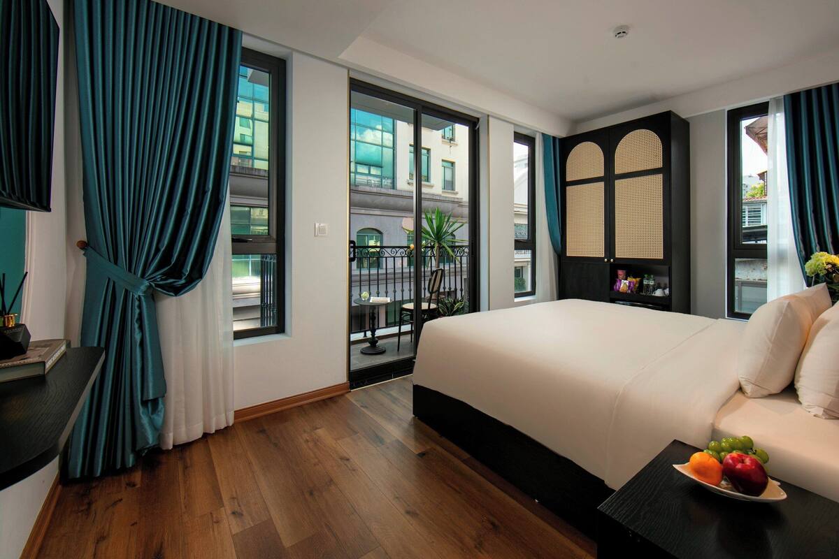 A hotel room with a large bed, a small table with fruit, and a view of the city through large windows.