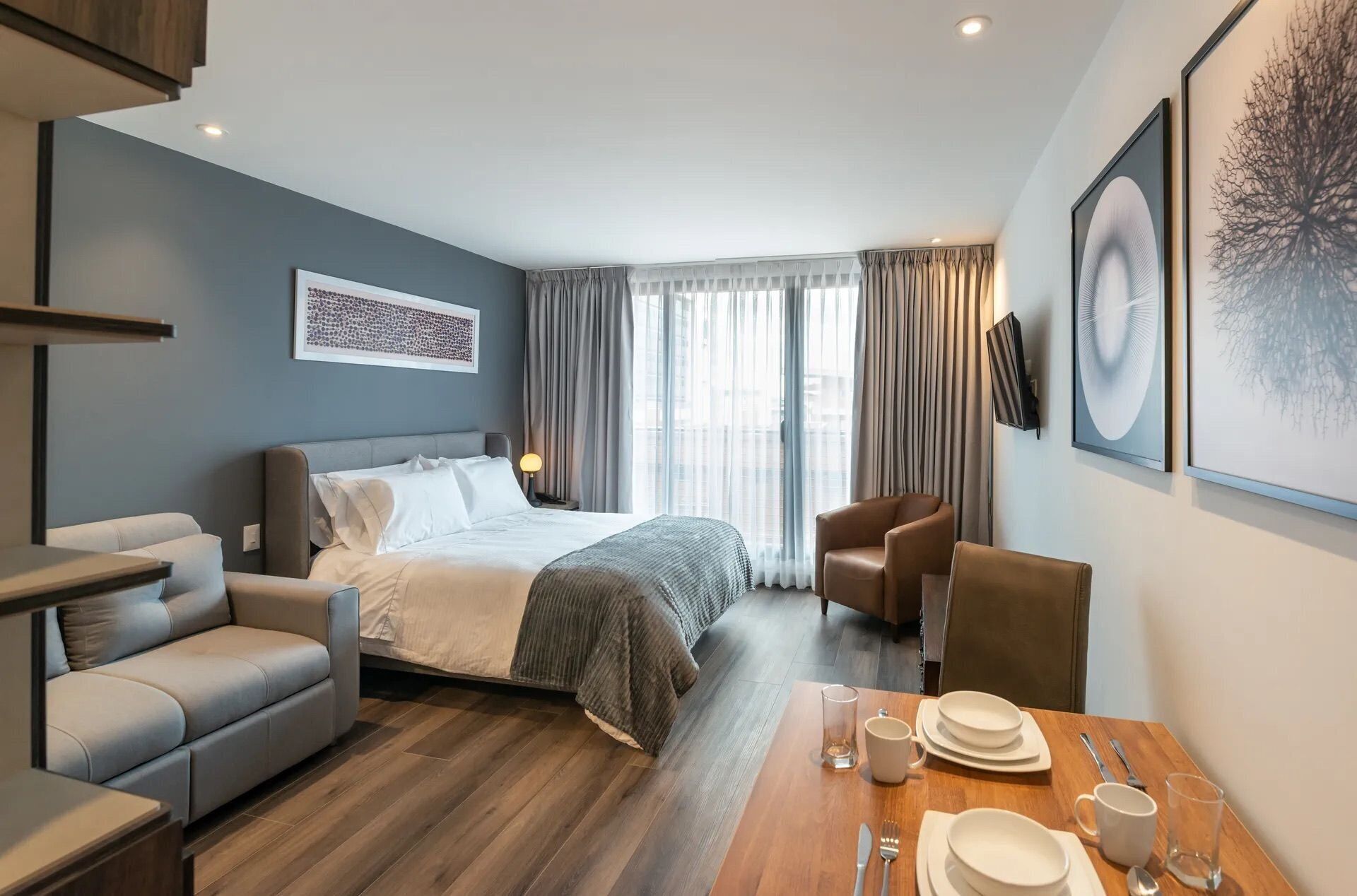 A modern hotel room with a bed, a sofa, a dining table set with plates, glasses, and cutlery.