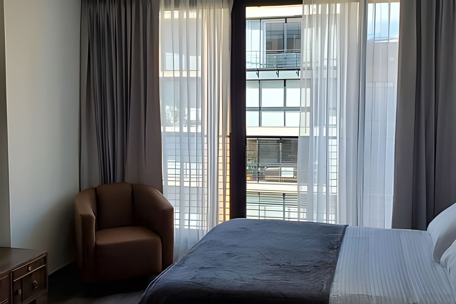 A hotel room with a bed, a chair, and large windows with sheer curtains.