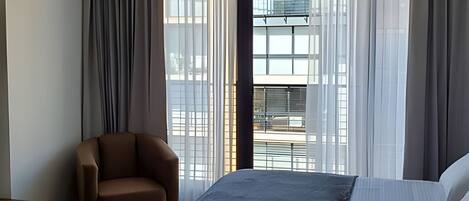 A hotel room with a bed, a chair, and large windows with sheer curtains.