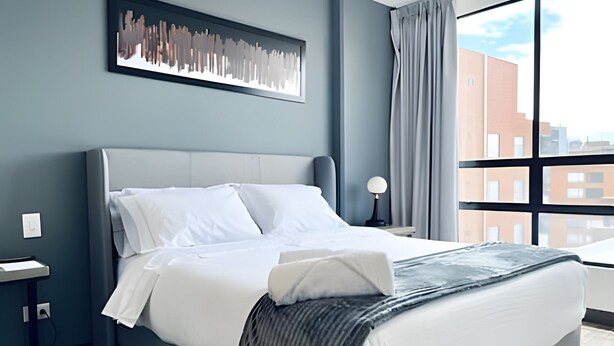 A modern hotel room with a large bed, a nightstand, a lamp, and a view of buildings through the window. - Morph Chico (Bogotá)