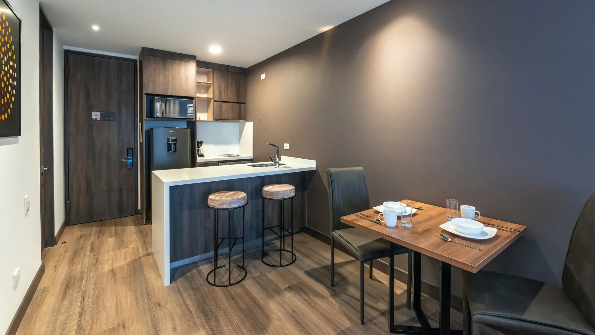 A compact kitchen with a central island, bar stools, a dining table set for two, and a refrigerator.
