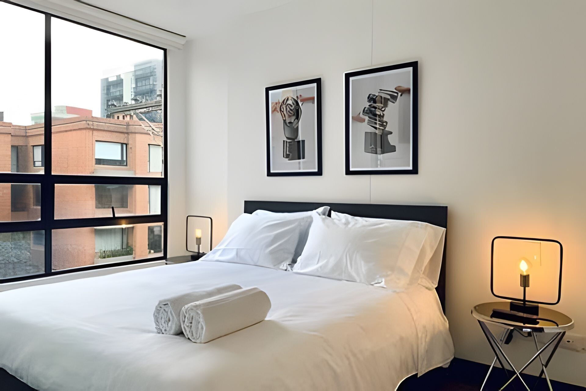 A hotel room with a large bed, two framed artworks on the wall, a bedside table with a lamp, and a window with a city view.