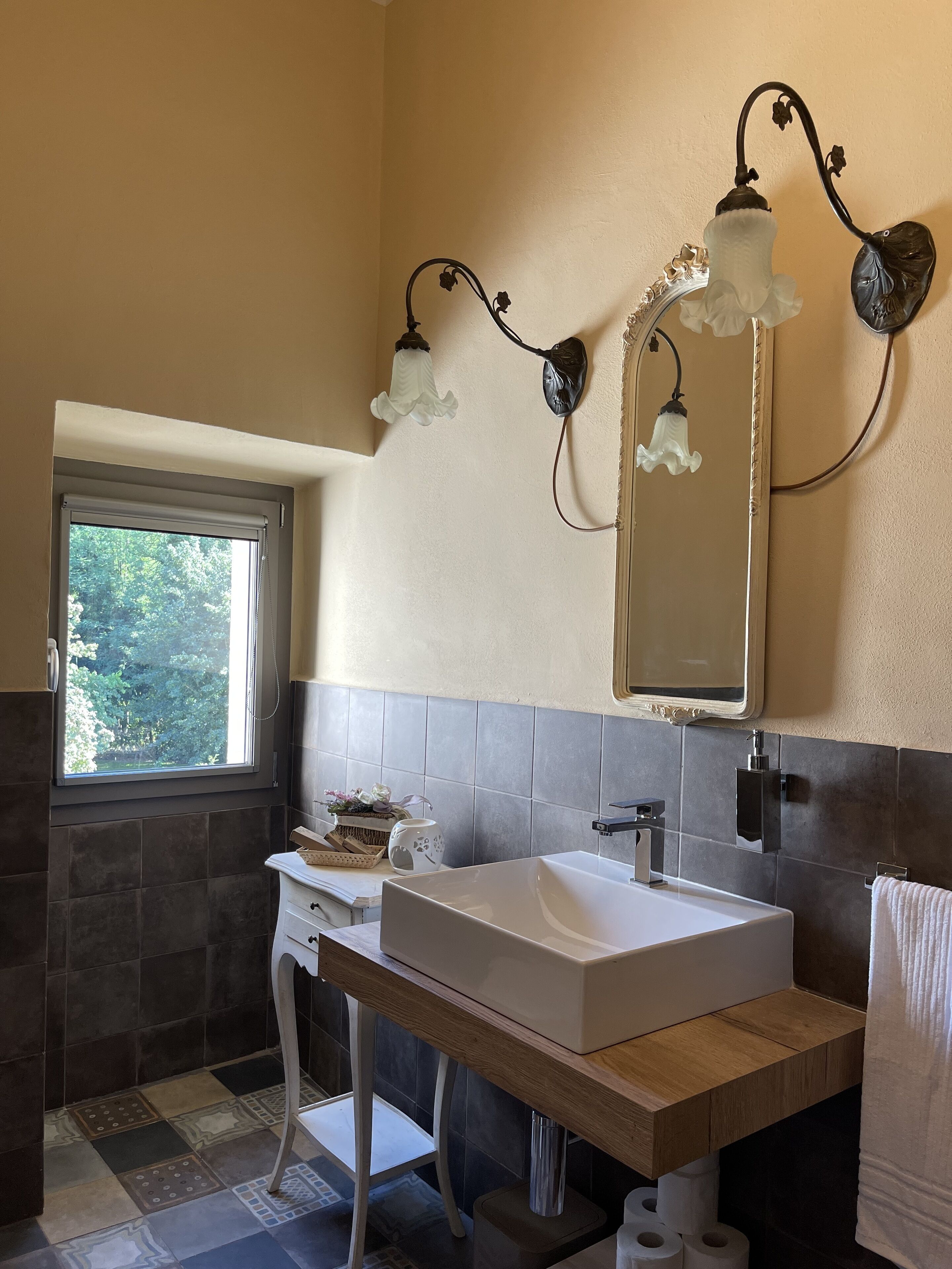 Une salle de bain dotée d’une grande fenêtre, d’un lavabo blanc, d’un miroir et de deux appliques murales.