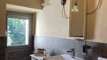 Une salle de bain dotée d’une grande fenêtre, d’un lavabo blanc, d’un miroir et de deux appliques murales.