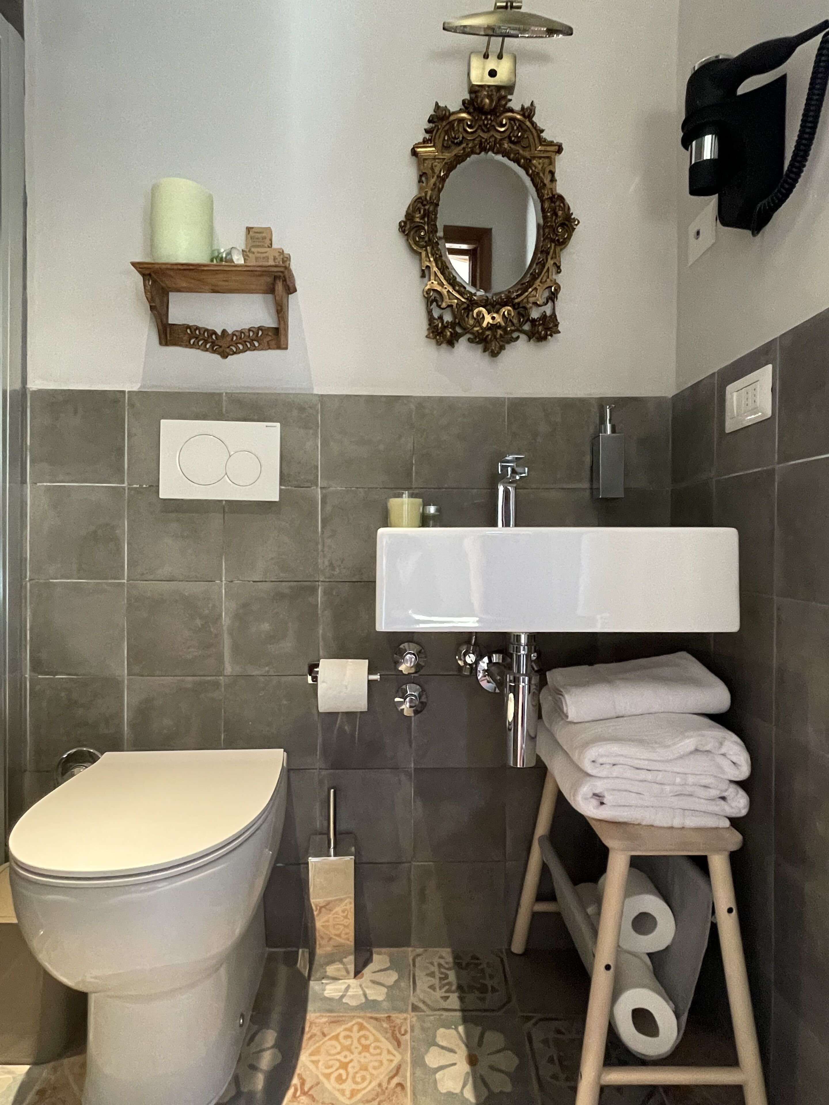 Ein Badezimmer mit Toilette, Waschbecken und Spiegel. Handtücher liegen auf einem hölzernen Hocker gestapelt.