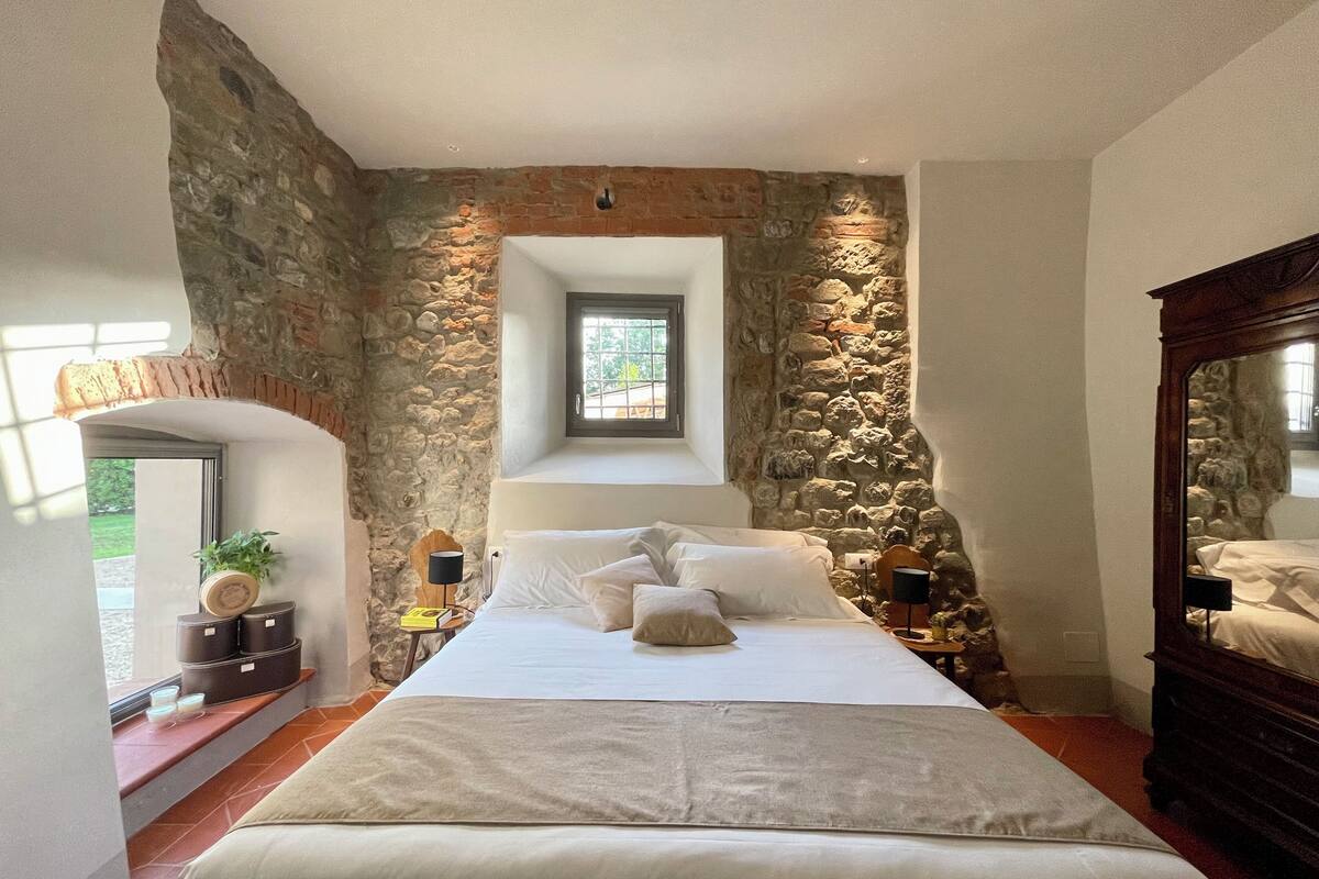 Un dormitorio con pared de piedra, una cama, una mesita de noche, un ventanal y un espejo.