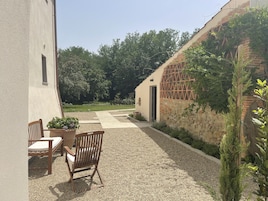 Un patio con un sendero de grava, sillas de madera y una mesa. Se observa un edificio con un muro de piedra y una puerta.