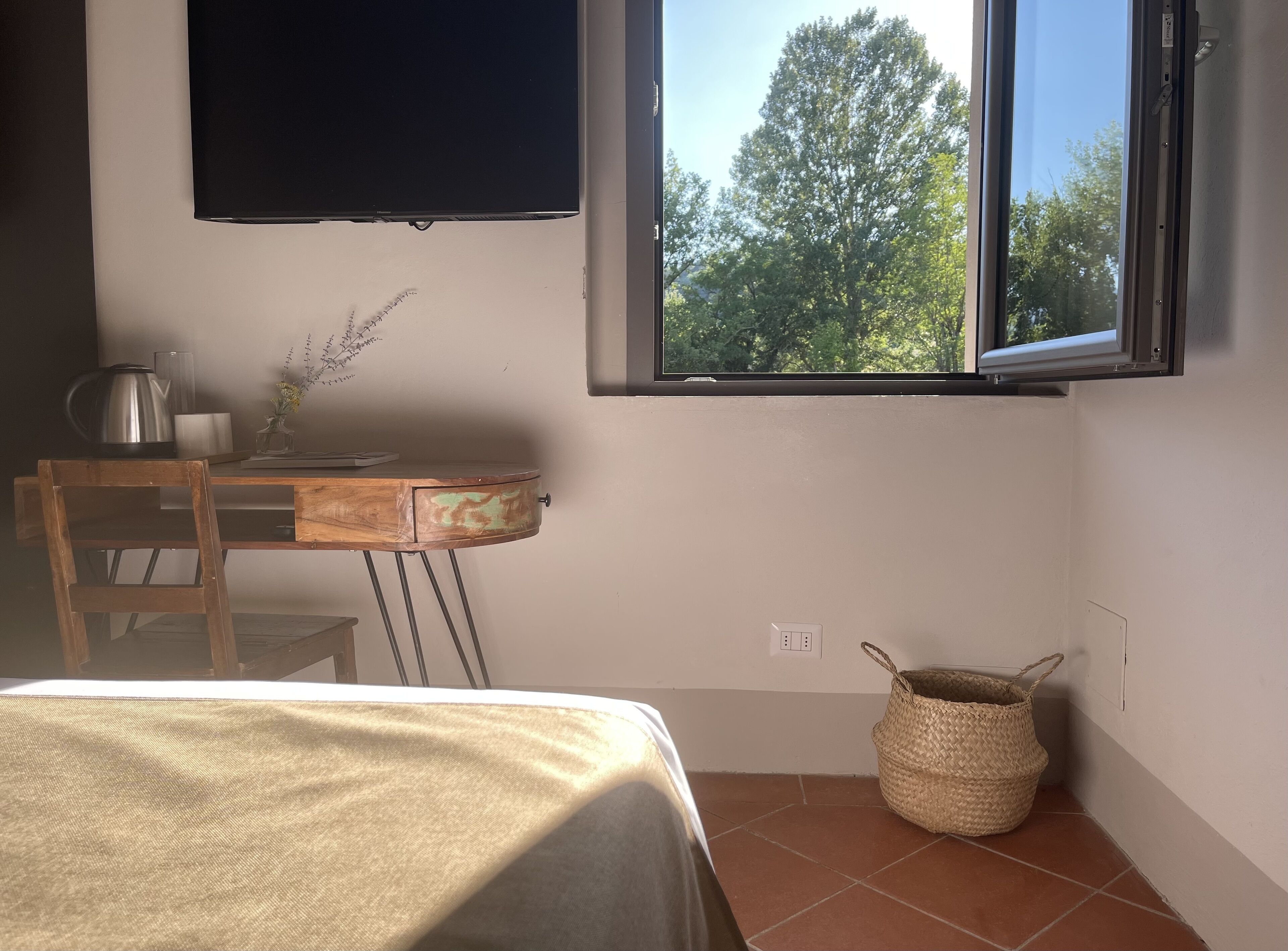 A room with a bed, a desk with a kettle, a lamp, a vase with flowers, a window with a view of trees, and a woven basket.