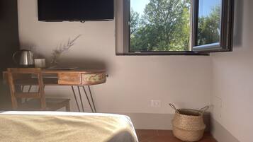 A room with a bed, a desk with a kettle, a lamp, a vase with flowers, a window with a view of trees, and a woven basket.