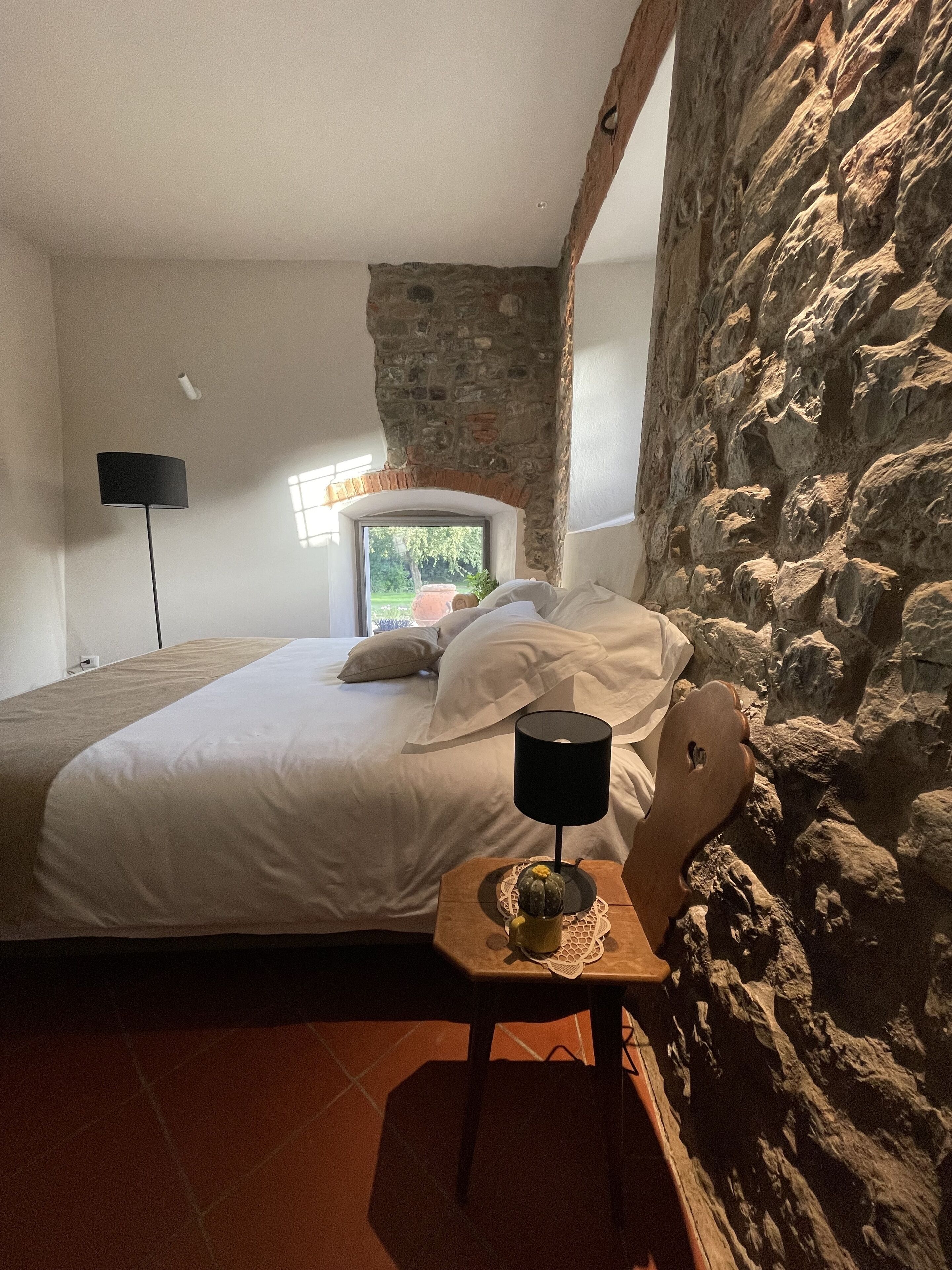 A bedroom with a stone wall, a bed with white linens, a nightstand with a lamp, and a window with a view of greenery.