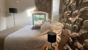A bedroom with a stone wall, a bed with white linens, a nightstand with a lamp, and a window with a view of greenery.