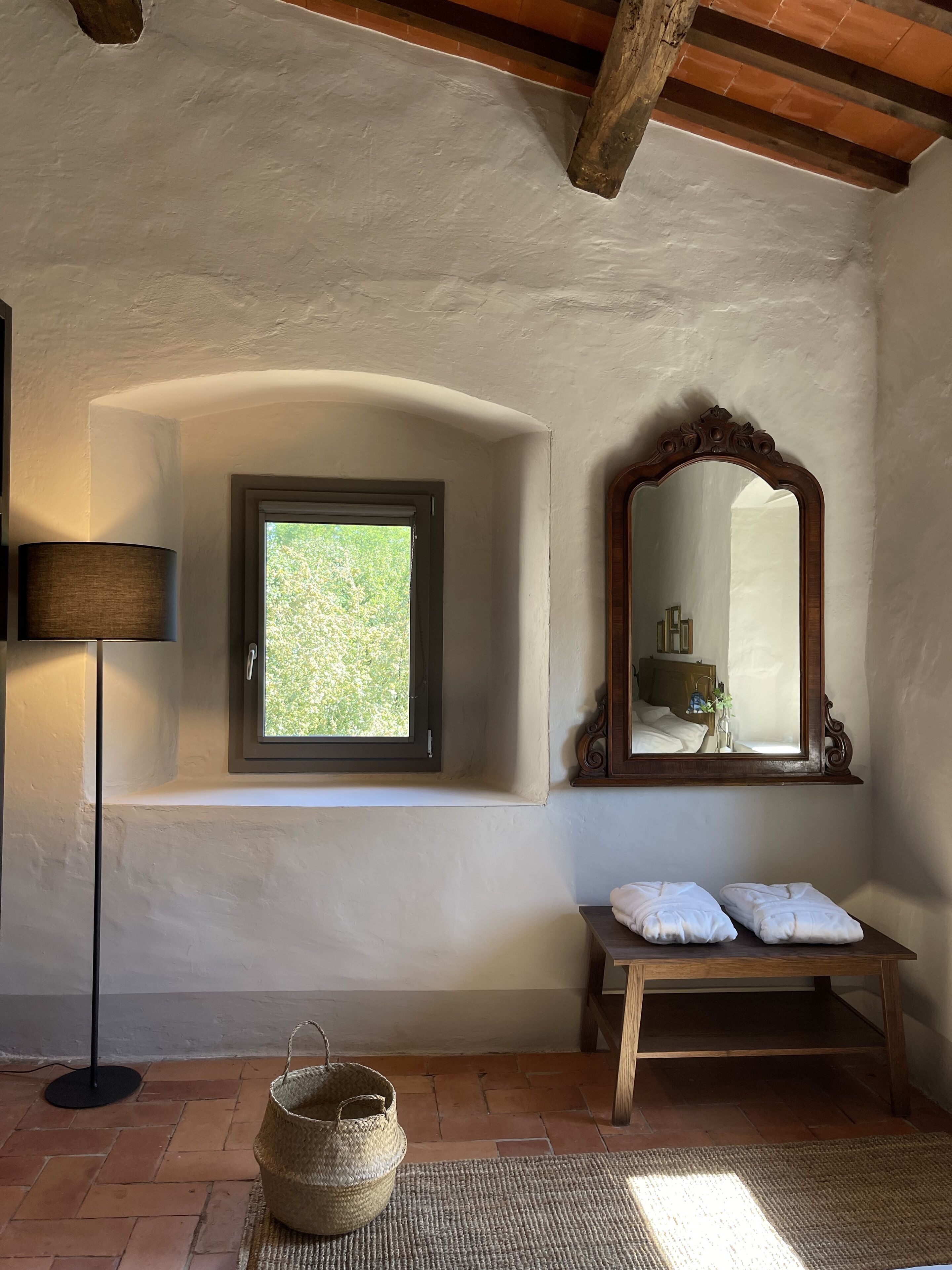 A room with a rustic ceiling, a window with a view of greenery, a wooden bench with towels, and a mirror with a wooden frame.