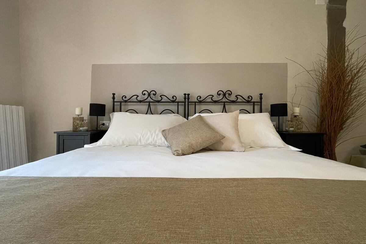 Una cama bien hecha con cabecera, dos mesitas de noche con lámparas y una planta alta junto a la cama.