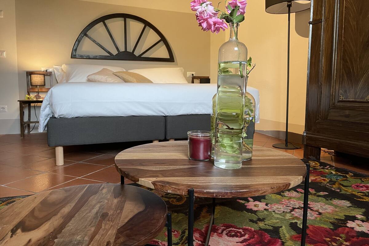 Un dormitorio con una cama, una mesita de noche, una lámpara, un florero con flores y una mesa con una vela.